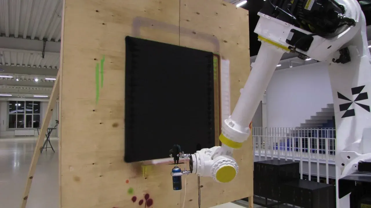 A six-axis robot arm sprays a black field onto a plywood wall